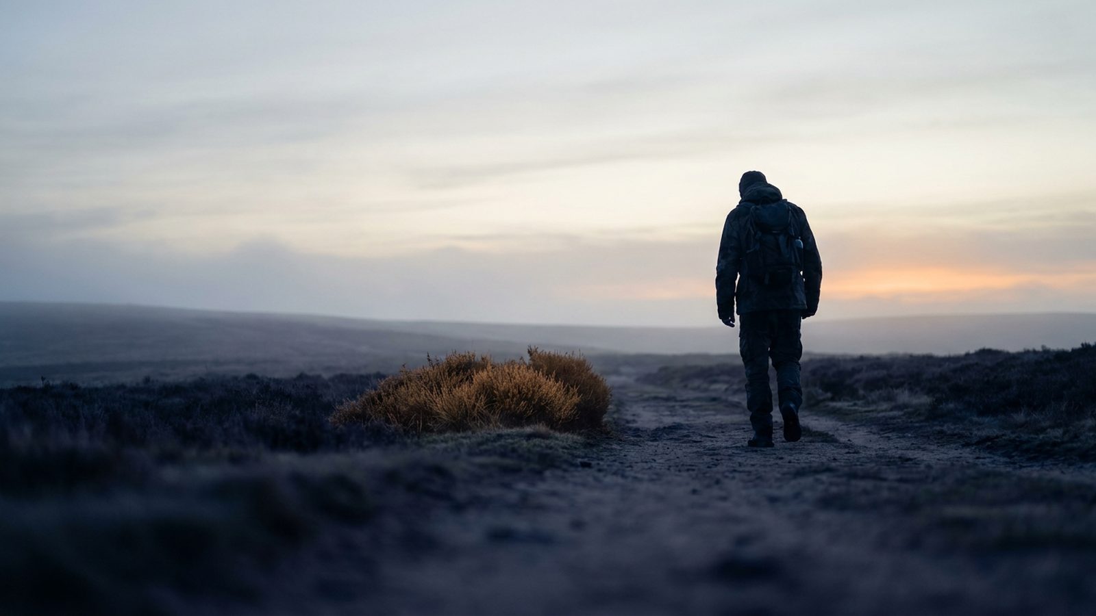 Person standing on a moorland path at dawn, silhouetted against a softly glowing sky.