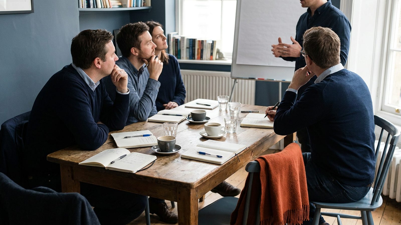 A small group of people in a meeting room are actively listening to one speaker, with notebooks on the table.