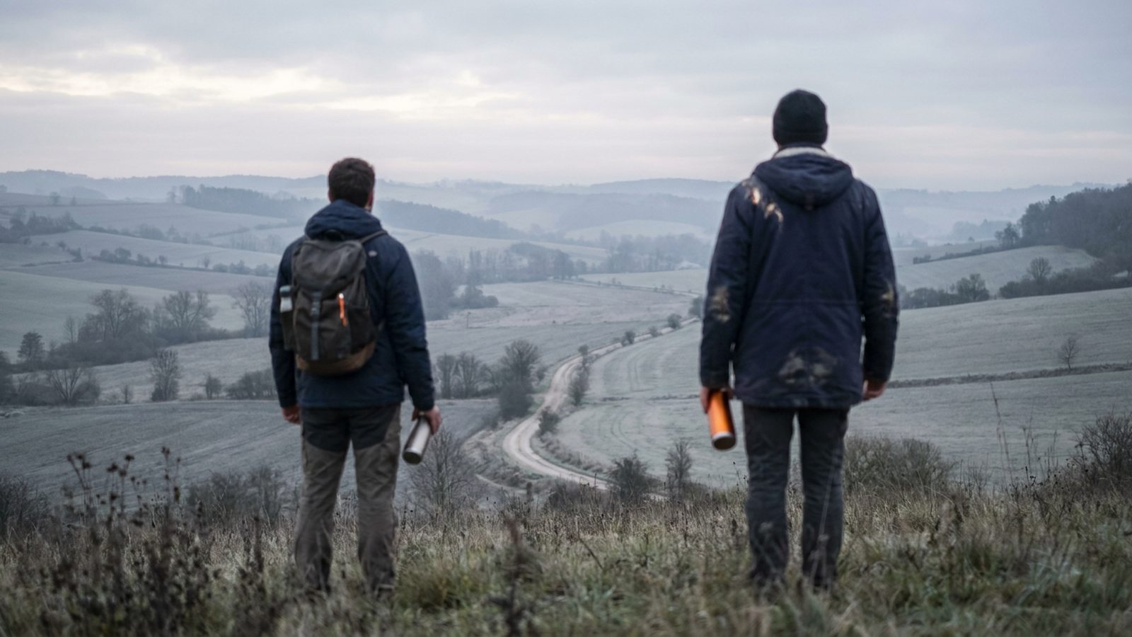 A lone hiker stands on a hill overlooking a peaceful, expansive rural landscape at dawn.